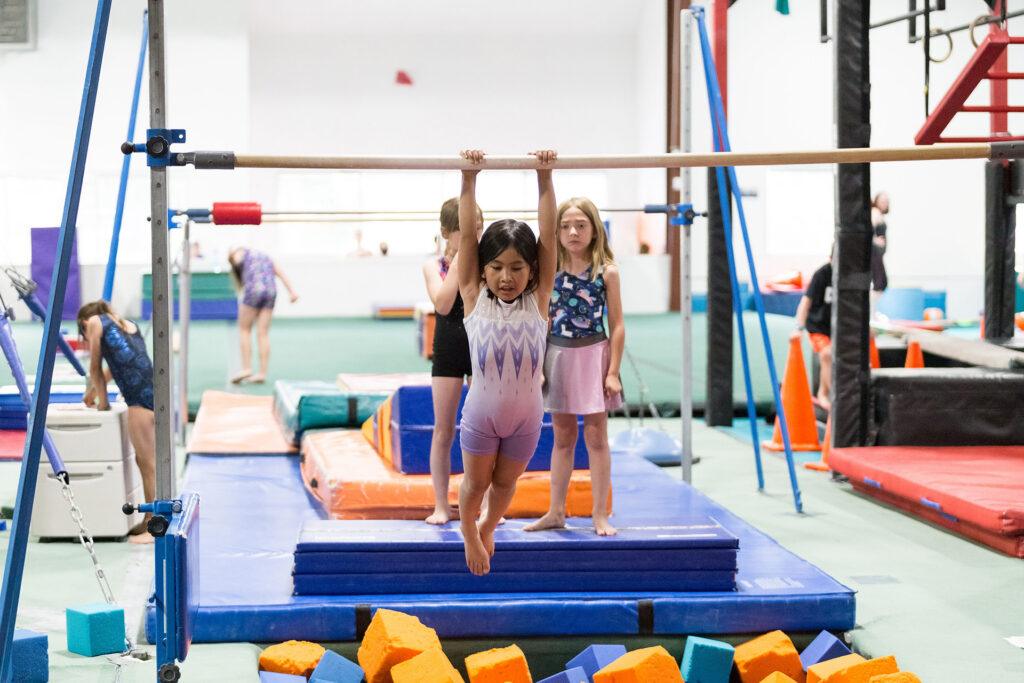 Young girl practicing gymnastics on the bars at Mountain Kids Louisville while other children wait their turn during an indoor gym class.
